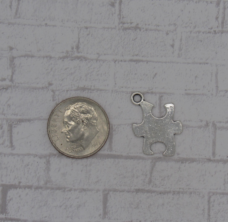 K&ouml;nnte beinhalten: Silberfarbener Metall-Puzzle-Anh&auml;nger mit einem Loch oben zum Anbringen an einer Halskette oder einem Armband. Der Anh&auml;nger ist etwa so gro&szlig; wie eine 10-Cent-M&uuml;nze.