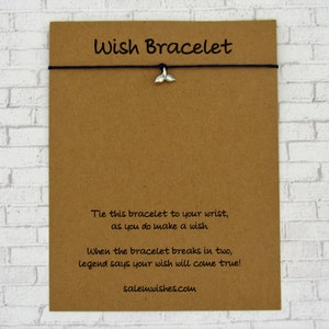 May include: A brown card with the text "Wish Bracelet" printed on it. The card has a black string with a silver whale tail charm attached to it. The text on the card reads: "Tie this bracelet to your wrist, as you do make a wish. When the bracelet breaks in two, legend says your wish will come true! salemwishes.com"