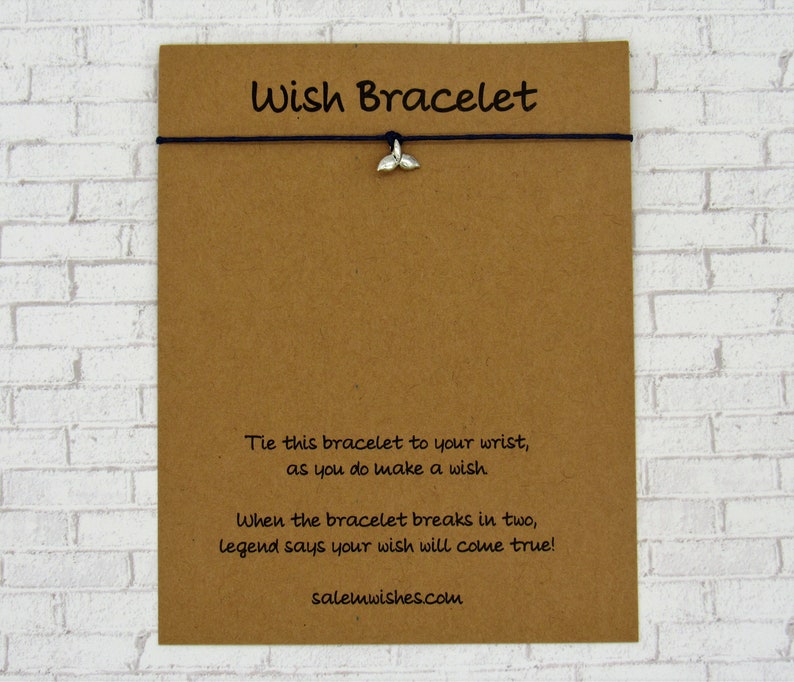 May include: A brown card with the text "Wish Bracelet" printed on it. The card has a black string with a silver whale tail charm attached to it. The text on the card reads: "Tie this bracelet to your wrist, as you do make a wish. When the bracelet breaks in two, legend says your wish will come true! salemwishes.com"