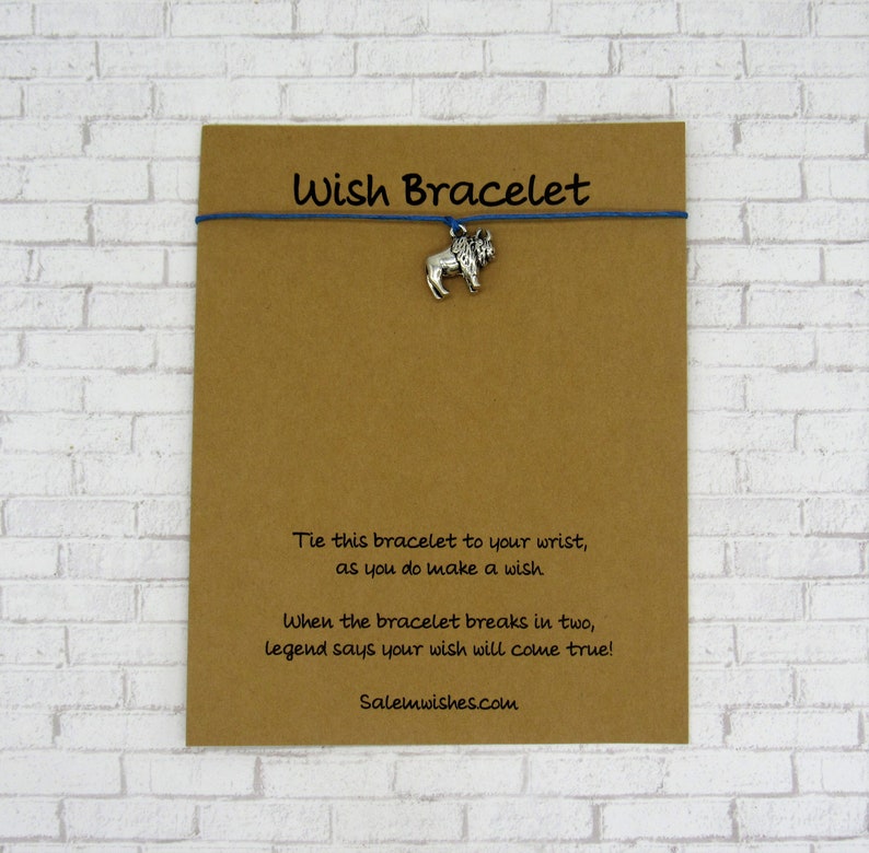 May include: A blue string bracelet featuring a small silver-toned lion charm attached to a brown card. The card displays the text Wish Bracelet, instructions to tie the bracelet while making a wish, and the website address Salemwishes.com.