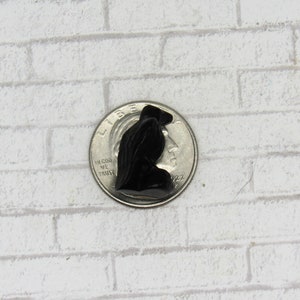 Pode incluir: Pequena figura preta esculpida de uma pessoa com uma capa com capuz, ao lado de uma moeda para escala. A figura est&aacute; sobre um fundo de tijolos brancos. A figura tem aproximadamente 2,5 cm de altura.
