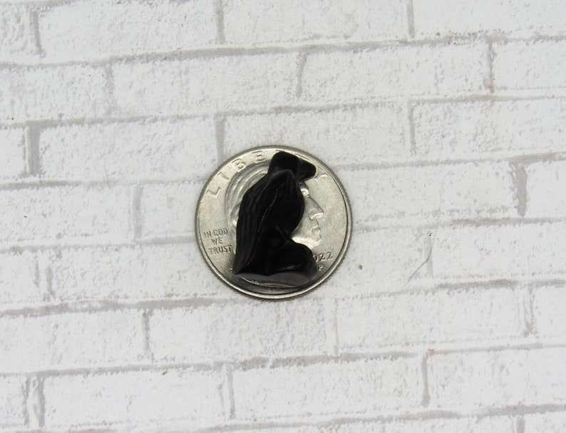 Pode incluir: Pequena figura preta esculpida de uma pessoa com uma capa com capuz, ao lado de uma moeda para escala. A figura est&aacute; sobre um fundo de tijolos brancos. A figura tem aproximadamente 2,5 cm de altura.