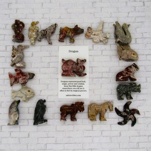 Op de afbeelding: Een verzameling kleine, handgesneden stenen dierenfiguren, waaronder een draak, een vis, een beer en een vogel. De figuren zijn gerangschikt op een witte bakstenen achtergrond met een kaart waarop "Drache" staat en een beschrijving van de betekenis van draken.