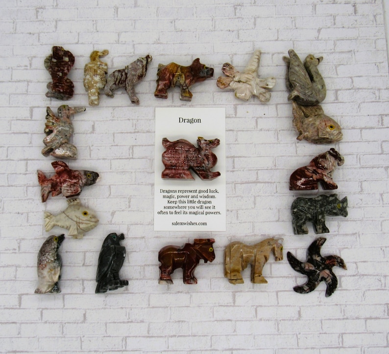 Op de afbeelding: Een verzameling kleine, handgesneden stenen dierenfiguren, waaronder een draak, een vis, een beer en een vogel. De figuren zijn gerangschikt op een witte bakstenen achtergrond met een kaart waarop "Drache" staat en een beschrijving van de betekenis van draken.