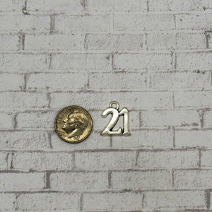 May include: A silver-colored charm with the number "21" next to a U.S. dime. The charm has a small loop at the top for attaching to a necklace or bracelet. The background is a white brick wall.