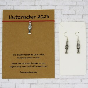 May include: A red string bracelet with a silver nutcracker charm. The bracelet is tied to a brown card with the text "Nutcracker 2023" and instructions on how to make a wish. The card also includes the website "Salemwishes.com".