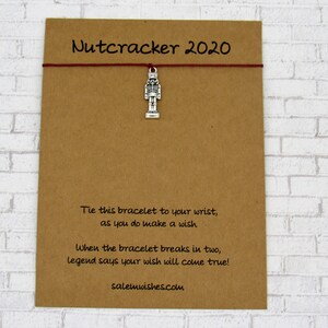 May include: A brown card with the text "Nutcracker 2020" and instructions for a wish bracelet. The card has a silver nutcracker charm attached to it with a red string.