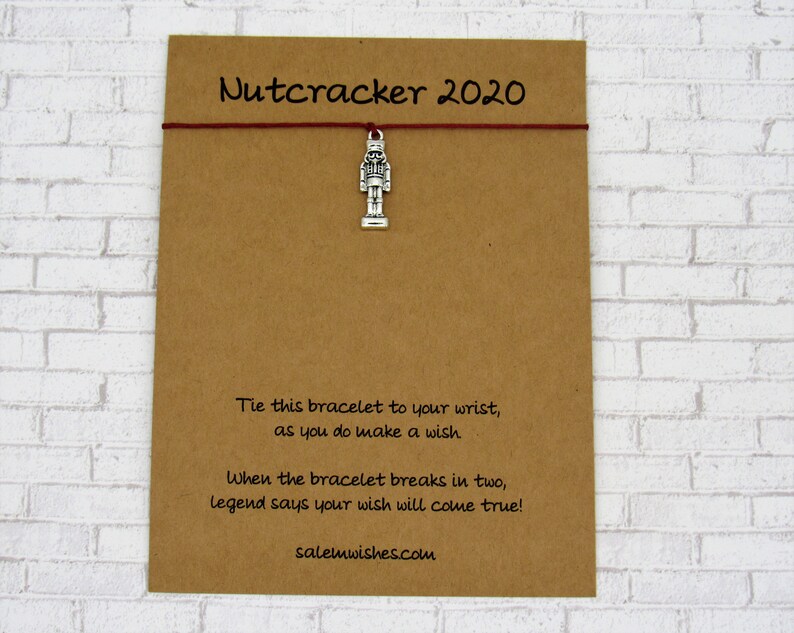 May include: A brown card with the text "Nutcracker 2020" and instructions for a wish bracelet. The card has a silver nutcracker charm attached to it with a red string.