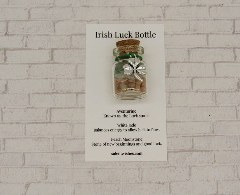May include: A small glass bottle with a cork stopper filled with green, white, and pink stones and a silver four-leaf clover charm. The bottle is labeled "Irish Luck Bottle" and includes a description of the stones: Aventurine, White Jade, and Peach Moonstone. The text also includes the website salemwishes.com.