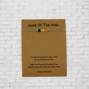 K&ouml;nnte beinhalten: Eine braune Karte mit dem Text "Luck Of The Irish" darauf gedruckt. Ein wei&szlig;es Armband aus Schnur mit gr&uuml;nen, wei&szlig;en und orangefarbenen Perlen ist an der Karte befestigt. Die Karte enth&auml;lt auch Anweisungen, wie man mit dem Armband einen Wunsch macht.