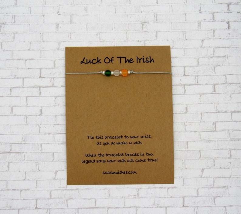 K&ouml;nnte beinhalten: Eine braune Karte mit dem Text "Luck Of The Irish" darauf gedruckt. Ein wei&szlig;es Armband aus Schnur mit gr&uuml;nen, wei&szlig;en und orangefarbenen Perlen ist an der Karte befestigt. Die Karte enth&auml;lt auch Anweisungen, wie man mit dem Armband einen Wunsch macht.