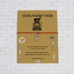 May include: A red string bracelet attached to a tan card featuring the text Nutcracker Wish and a nutcracker illustration. The card displays silver-toned charms including an angel, a nutcracker, a crown, a fairy, a ballet slipper, and a candy cane.
