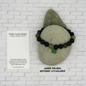 K&ouml;nnte beinhalten: Schwarzes und gr&uuml;nes Perlenarmband mit einem goldenen Kleeblatt-Anh&auml;nger. Das Armband liegt auf einem grauen Stein mit einer wei&szlig;en Karte, auf der "Irish Luck Stone Abundance, Happiness and Luck" und "Wear this natural crystal bracelets to benefit from the healing powers or when you need a little Irish Luck. www.salemwishes.com" steht. Die Karte tr&auml;gt au&szlig;erdem den Text "More Charm Options Available".