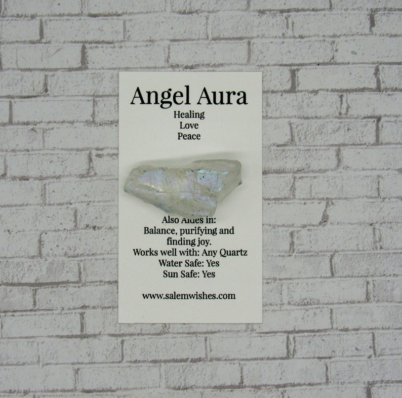 May include: A white Angel Aura crystal with iridescent shimmer. The card reads "Angel Aura", "Healing", "Love", "Peace", "Also Aues in: Balance, purifying and finding joy.", "Works well with: Any Quartz", "Water Safe: Yes", "Sun Safe: Yes", "www.salemwishes.com".