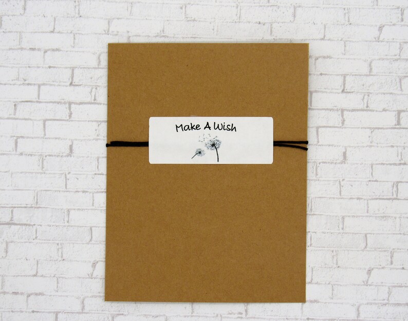 May include: A brown paper envelope with a white label that says "Make A Wish" and has a black and white illustration of dandelions. The envelope is tied with a black string.