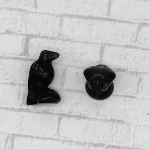 Pode incluir: Duas estatuetas de corvo pretas. Um corvo est&aacute; em p&eacute;, e o outro &eacute; uma vista da cabe&ccedil;a e do peito. As estatuetas s&atilde;o feitas de um material escuro e brilhante, sobre um fundo de tijolos brancos.
