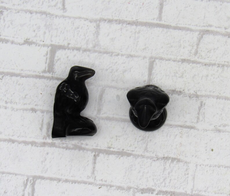 Pode incluir: Duas estatuetas de corvo pretas. Um corvo est&aacute; em p&eacute;, e o outro &eacute; uma vista da cabe&ccedil;a e do peito. As estatuetas s&atilde;o feitas de um material escuro e brilhante, sobre um fundo de tijolos brancos.