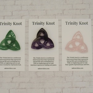 Peut inclure: Trois nœuds de la Trinité, chacun fait d'un type de pierre différent. Le vert est en aventurine, le violet en améthyste et le rose en quartz rose. Chaque nœud est sur une carte blanche avec le texte "Trinity Knot" et une description du nœud et de la pierre.