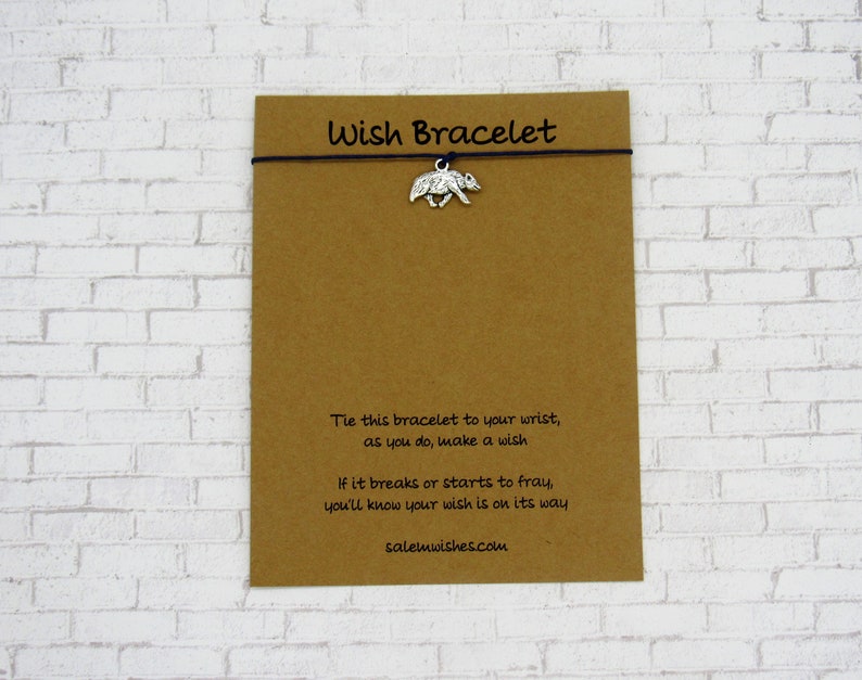 May include: A brown card with the text "Wish Bracelet" printed on it. The card has a black string with a silver charm of a bear attached to it. The text on the card reads: "Tie this bracelet to your wrist, as you do, make a wish. If it breaks or starts to fray, you'll know your wish is on its way. salemwishes.com"