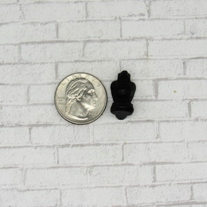 Pode incluir: Um pequeno objeto decorativo preto com uma forma &uacute;nica, possivelmente uma escultura ou um amuleto em miniatura. O objeto est&aacute; posicionado ao lado de uma moeda de um quarto de d&oacute;lar dos EUA para compara&ccedil;&atilde;o de tamanho. O fundo &eacute; um padr&atilde;o de tijolos brancos.