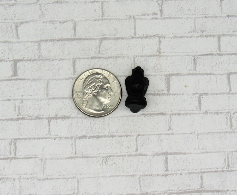 Pode incluir: Um pequeno objeto decorativo preto com uma forma &uacute;nica, possivelmente uma escultura ou um amuleto em miniatura. O objeto est&aacute; posicionado ao lado de uma moeda de um quarto de d&oacute;lar dos EUA para compara&ccedil;&atilde;o de tamanho. O fundo &eacute; um padr&atilde;o de tijolos brancos.