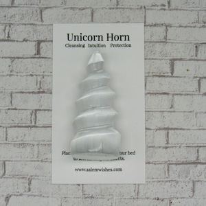 Op de afbeelding: Wit selenietkristal in de vorm van een eenhoornhoorn. De kaart luidt "E&eacute;nhoornhoorn Reiniging Intu&iuml;tie Bescherming Plaats in de buurt van je bed of voorwerpen. www.salemwishes.com"