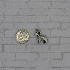 May include: A silver-toned metal charm shaped like a ram with curled horns. The charm is about the size of a US dime.