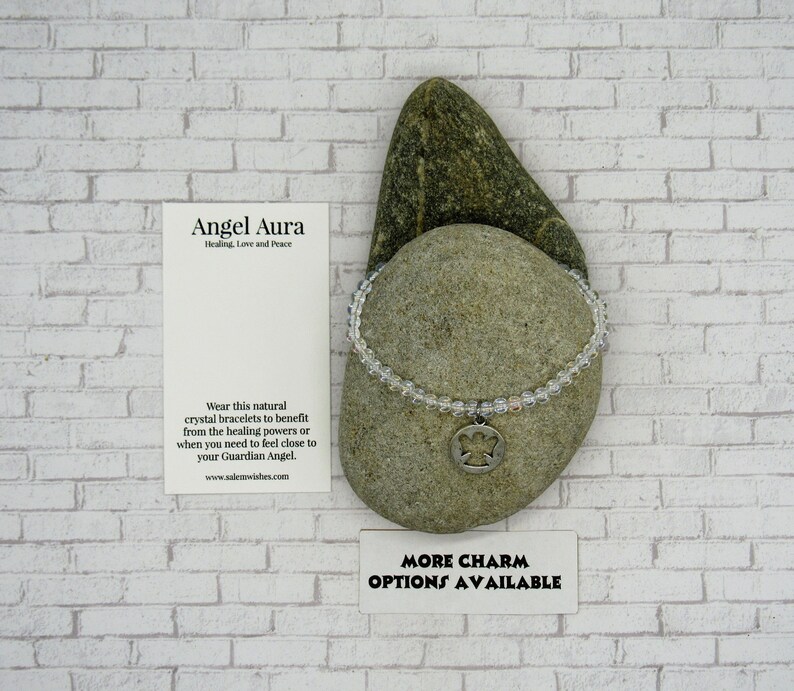 May include: A clear crystal bracelet with a silver angel charm. The bracelet is on a gray rock with a white brick background. The text on the card reads "Angel Aura Healing, Love and Peace" and "Wear this natural crystal bracelets to benefit from the healing powers or when you need to feel close to your Guardian Angel. www.salemwishes.com". The text on the bottom card reads "MORE CHARM OPTIONS AVAILABLE".