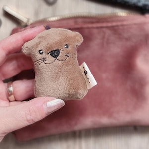 Könnte beinhalten: Ein kleines, plüschiges Otterspielzeug in Brauntönen, mit gestickten Gesichtszügen und einem kleinen Etikett. Der Otter wird vor einem rosa Beutel gehalten, was einen weichen, strukturierten Kontrast erzeugt.