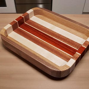 Op de afbeelding: Een rechthoekige houten dienblad met een verzonken interieur. De lade heeft afwisselende strepen van licht en donker hout, waaronder rode en natuurlijke tinten. De hoeken zijn afgerond en de lade is ongeveer 30 cm lang en 25 cm breed.