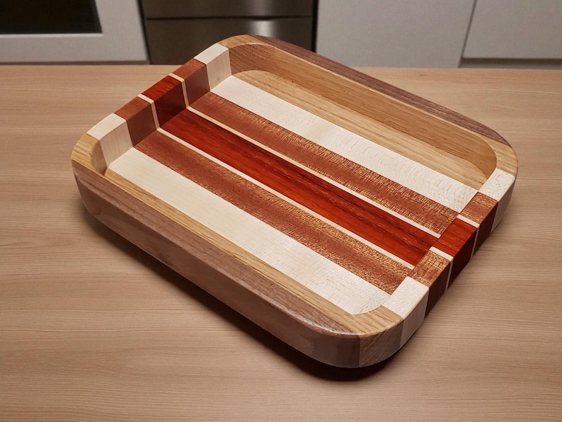 Op de afbeelding: Een rechthoekige houten dienblad met een verzonken interieur. De lade heeft afwisselende strepen van licht en donker hout, waaronder rode en natuurlijke tinten. De hoeken zijn afgerond en de lade is ongeveer 30 cm lang en 25 cm breed.