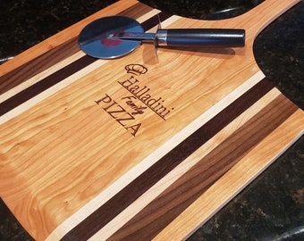 Op de afbeelding: Een houten pizzabord met een handvat, met een multi-houten ontwerp met strepen van kers, esdoorn en walnoot. Het bord is gegraveerd met de tekst "Halladimi Family Pizza". Een pizzames is op het bord geplaatst.