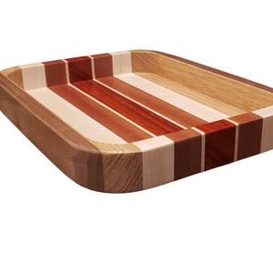 Op de afbeelding: Een rechthoekige houten dienblad met een gestreept ontwerp. De lade heeft afwisselende strepen van lichte en donkere houttinten, waaronder rood, bruin en beige. De randen zijn afgerond en de lade is gemaakt van verschillende houtsoorten.