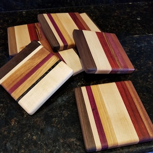 May include: Six square-shaped wooden cutting boards with a striped pattern of different wood types, including maple, walnut, and cherry. The boards are stacked on top of each other on a black countertop.