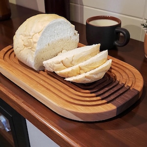 Puede incluir: Una hogaza de pan rebanado descansa sobre una tabla de pan de madera con un diseño de dos tonos. La tabla tiene un color de madera clara con un acento de madera más oscura. Una taza oscura y una pequeña planta en maceta también están sobre la mesa.