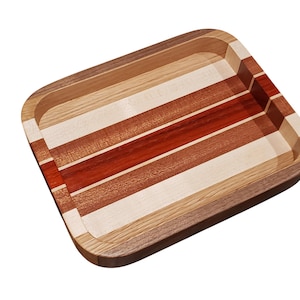 Op de afbeelding: Rechthoekige houten dienblad met afgeronde hoeken. De lade heeft afwisselende strepen van licht en donker hout, waaronder rode, bruine en natuurlijke tinten. De randen zijn van donkerder hout, waardoor een duidelijke rand ontstaat.