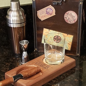 Peut inclure: Un porte-cigare et &agrave; boisson en bois avec le nom personnalis&eacute; "CHARLES" grav&eacute; sur la surface. Le porte-cigare a deux indentations circulaires, l'une pour un cigare et l'autre pour un verre. Un cigare repose dans l'indentation du cigare.