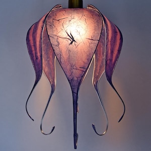 Può includere: Un'esclusiva lampada a sospensione fatta a mano. Il paralume presenta un design a strati con una combinazione di colori viola e rosa. La luce è realizzata in un materiale strutturato e ha un telaio in metallo. Il tema è floreale o botanico.
