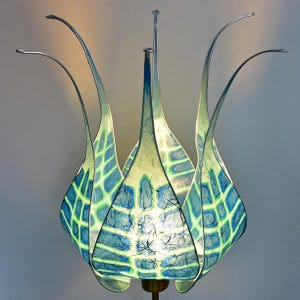 Könnte beinhalten: Eine handgefertigte dekorative Lampe mit floralem Design. Die Lampe hat einen blau-grün gemusterten Lampenschirm, der von schlanken, gebogenen Metallarmen getragen wird. Der Schirm hat ein gitterartiges Muster und die Lampe strahlt ein weiches, warmes Licht aus.