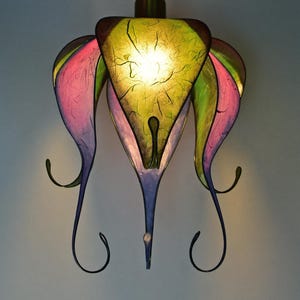 Può includere: Una lampada a sospensione colorata e realizzata a mano. La lampada presenta un paralume centrale triangolare giallo e verde, circondato da paralumi a forma di petalo rosa, viola e verde. Ha uno stelo in metallo e dettagli decorativi in metallo curvo.