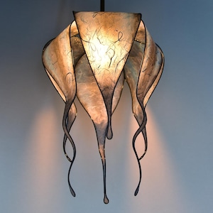 Könnte beinhalten: Eine einzigartige, handgefertigte Pendelleuchte. Die Leuchte hat einen strukturierten, durchscheinenden Schirm mit einem geschichteten, organischen Design. Der Schirm wird von einem dunklen Metallrahmen getragen, mit fließenden, länglichen Akzenten, die einen dramatischen Effekt erzeugen. Das Licht wird von innen beleuchtet.