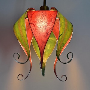 Könnte beinhalten: Eine handgefertigte Pendelleuchte mit einem einzigartigen floralen Design. Der Schirm hat strukturiertes Papier in Rot, Orange und Grün. Die Leuchte hat dekorative Metallverzierungen und einen dunkelbraunen Stiel.