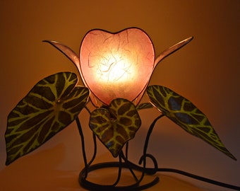 May include: A table lamp with a pink heart-shaped shade and three green leaves. The lamp is made of metal and has a black cord.