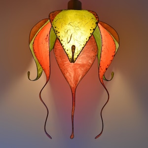 Könnte beinhalten: Ein farbenfroher, handgefertigter Lampenschirm in Form einer Blume mit rosa, grünen und gelben Blütenblättern. Der Lampenschirm ist aus Papier gefertigt und hat ein zartes, fließendes Design.