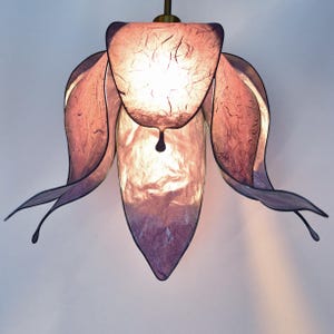Puede incluir: Una lámpara colgante decorativa con forma de flor. La lámpara presenta pétalos en capas en tonos morados y rosas. La fuente de luz es visible a través del material translúcido, creando un brillo suave. La lámpara está suspendida de un tallo color latón.