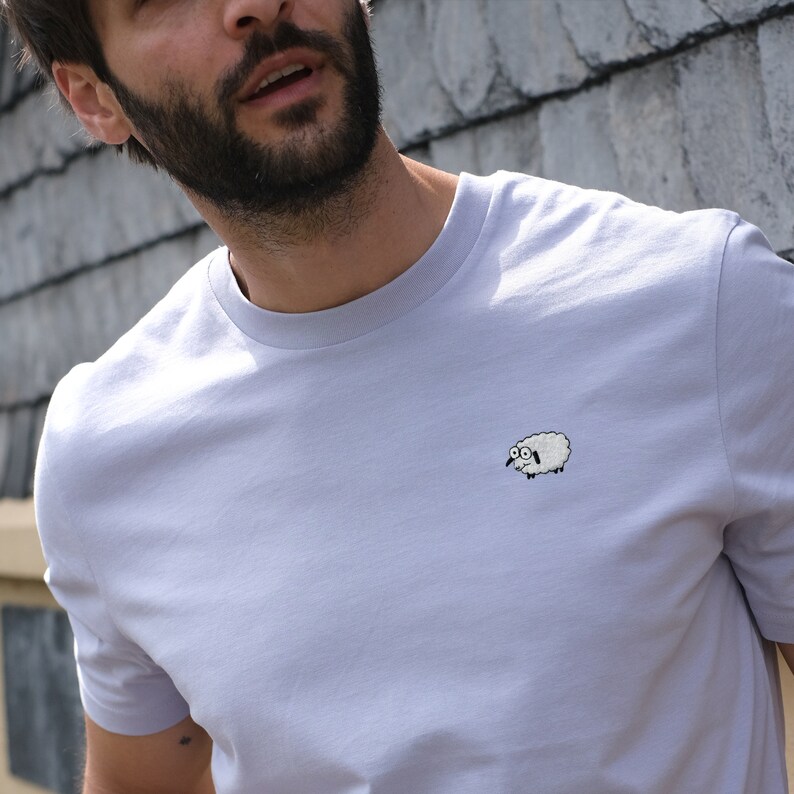 K&ouml;nnte beinhalten: Helllila T-Shirt mit einem kleinen, gestickten Schafmotiv auf der Brust. Das Schaf ist wei&szlig; mit schwarzen Details und einem &uuml;berraschten Gesichtsausdruck. Das T-Shirt hat kurze &Auml;rmel und einen Rundhalsausschnitt.