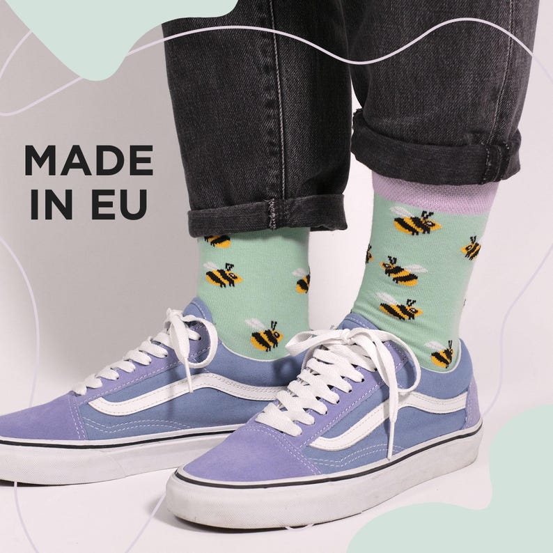 May include: Light blue trainers with white laces and soles. The person is wearing light green socks patterned with yellow and black bees. The text "MADE IN EU" is visible.