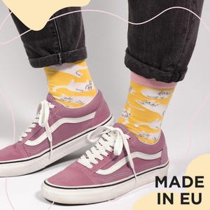 May include: A pair of pink sneakers with white laces and a white stripe on the side. The person is wearing yellow socks with a white cat pattern and dark gray jeans. The text "MADE IN EU" is visible.