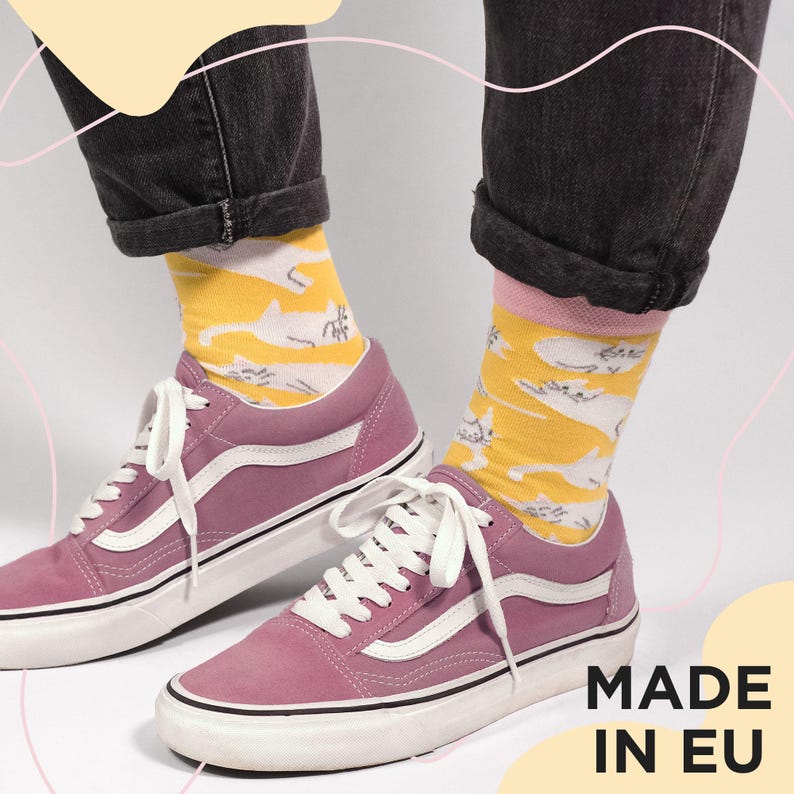May include: A pair of pink sneakers with white laces and a white stripe on the side. The person is wearing yellow socks with a white cat pattern and dark gray jeans. The text "MADE IN EU" is visible.