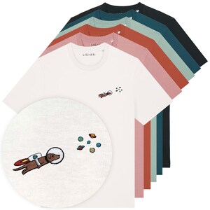 Può includere: Una pila di magliette a maniche corte in bianco, rosa, terracotta, verde salvia, verde acqua e nero. Ogni maglietta presenta un piccolo ricamo sul petto che mostra un cane marrone con casco spaziale e jetpack vicino a pianeti colorati.